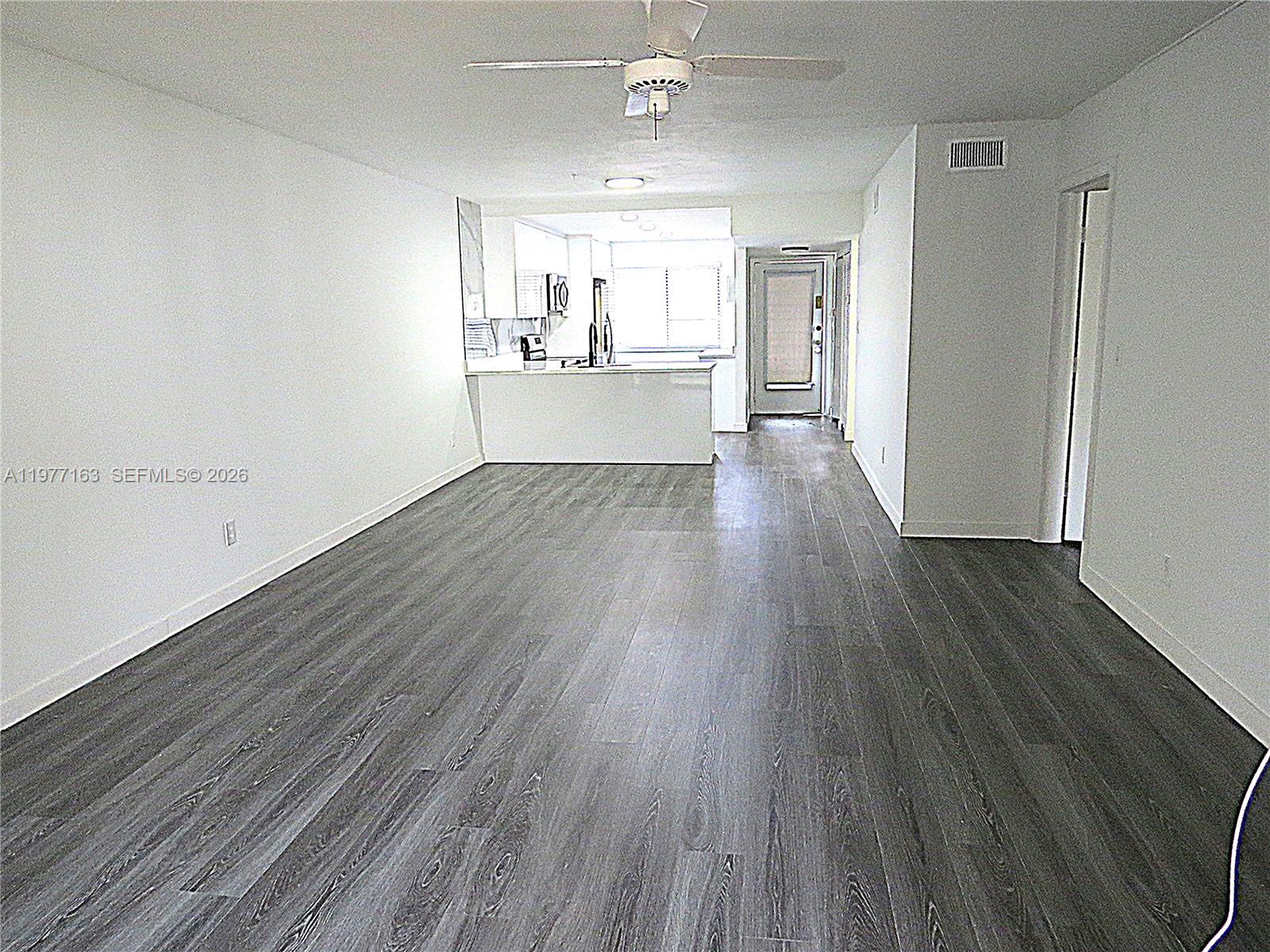 An open-concept living area features gray wood-look flooring, white walls, and a kitchen beyond — 3591 Inverrary Dr # 203, Lauderhill FL 33319, LAUDERHILL, FL