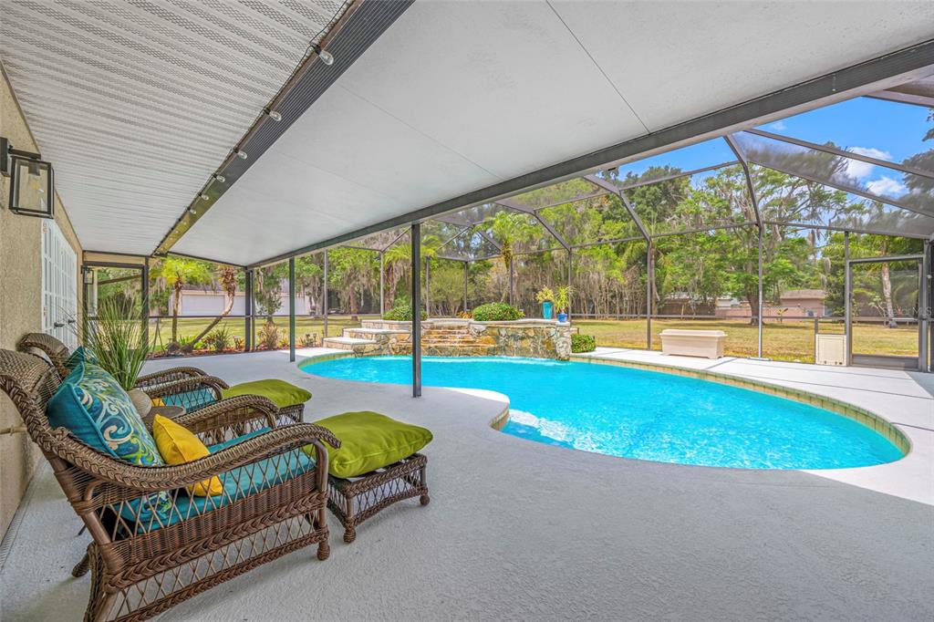 A covered patio with wicker furniture overlooks a swimming pool with a stone water feature — 15314 LAKE MAGDALENE BOULEVARD, TAMPA, FL