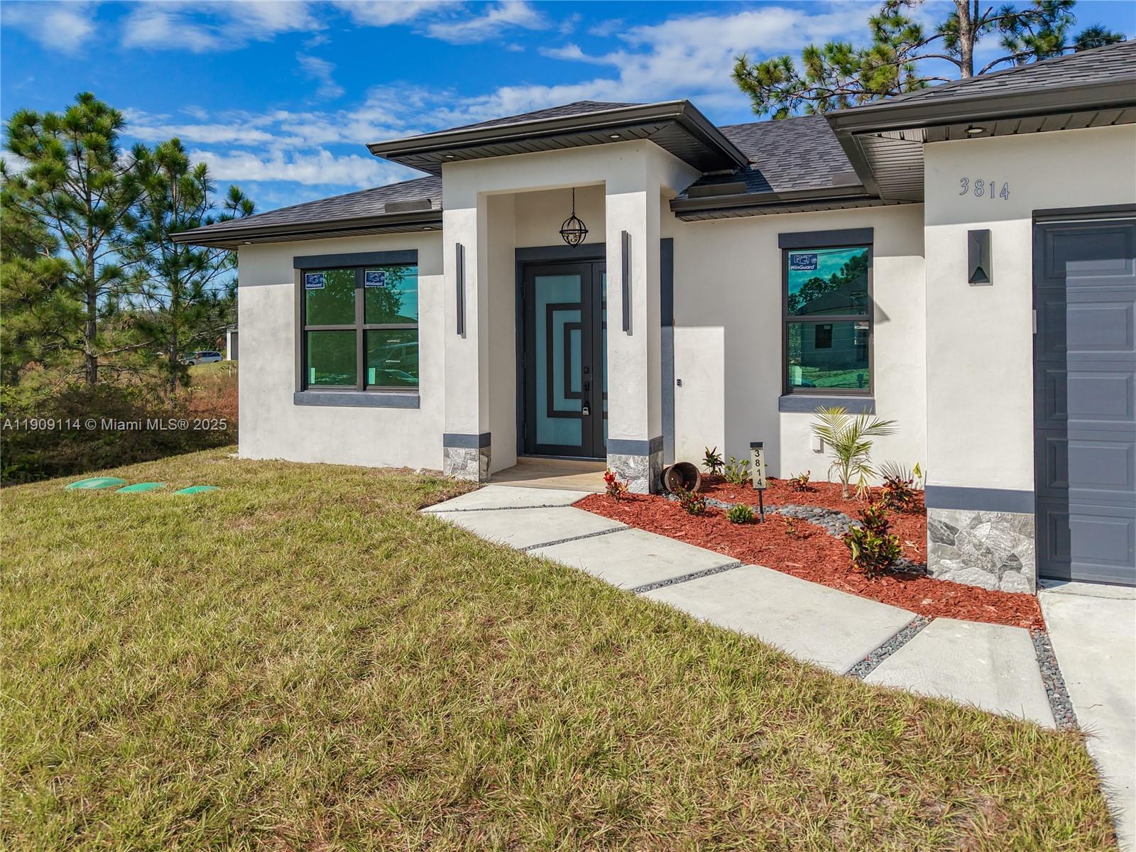 The exterior of a modern one-story house features white stucco walls, dark trim, and stone accents — 3814 9th St SW, Lehigh Acres FL 33976, LEHIGH ACRES, FL