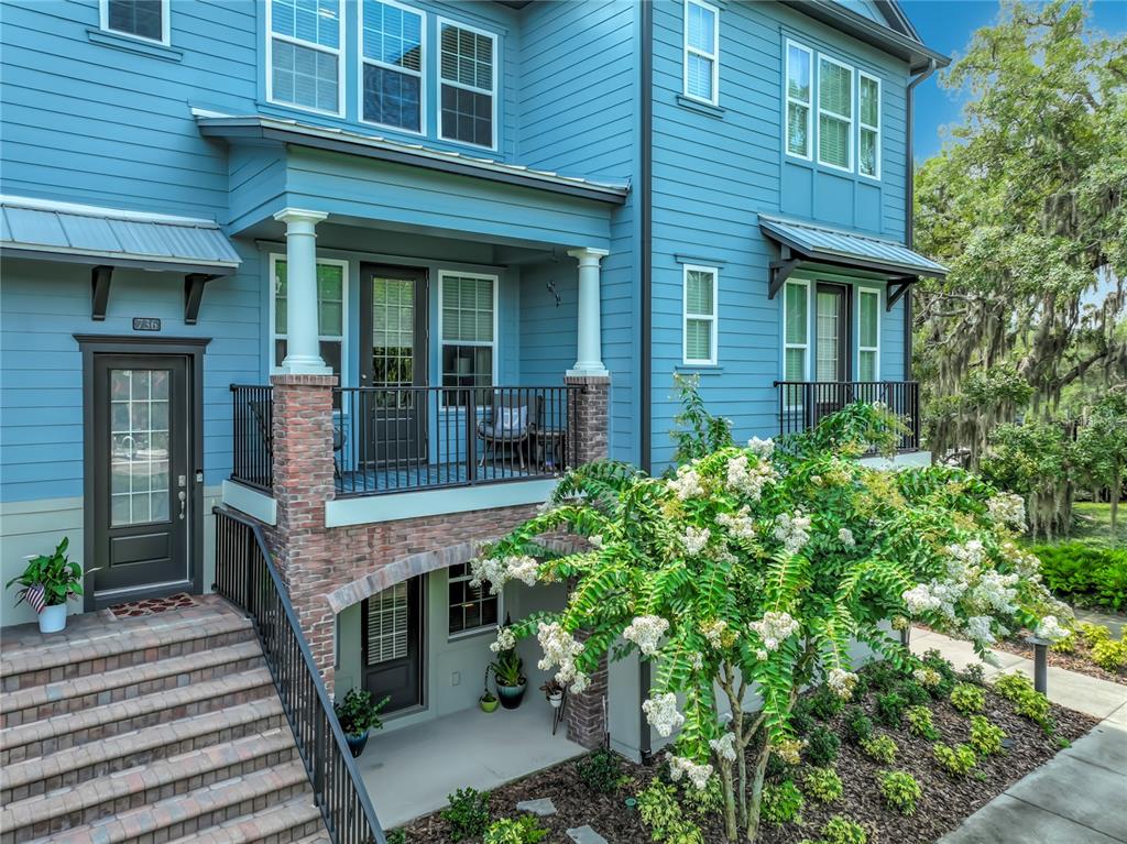 A blue multi-story house features a brick-accented porch, stairs, and a flowering tree — 736 CATALONIA COVE, OVIEDO, FL