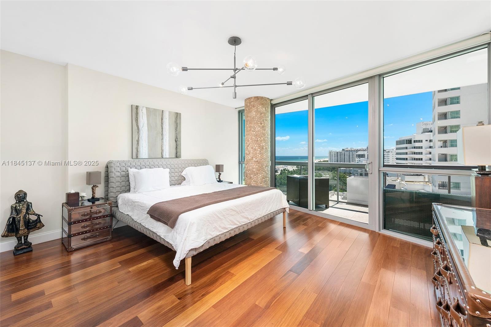 A bedroom features a bed, hardwood floors, a modern chandelier, and a balcony with ocean views — 101 20th St # 1706, Miami Beach FL 33139, MIAMI BEACH, FL