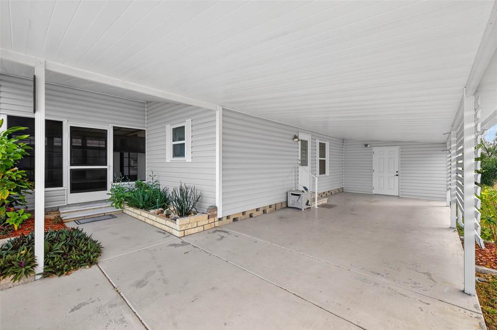 A covered concrete carport sits beside a light gray house with a screen door entrance — 3904 WILLIAM HUME DRIVE, ZEPHYRHILLS, FL