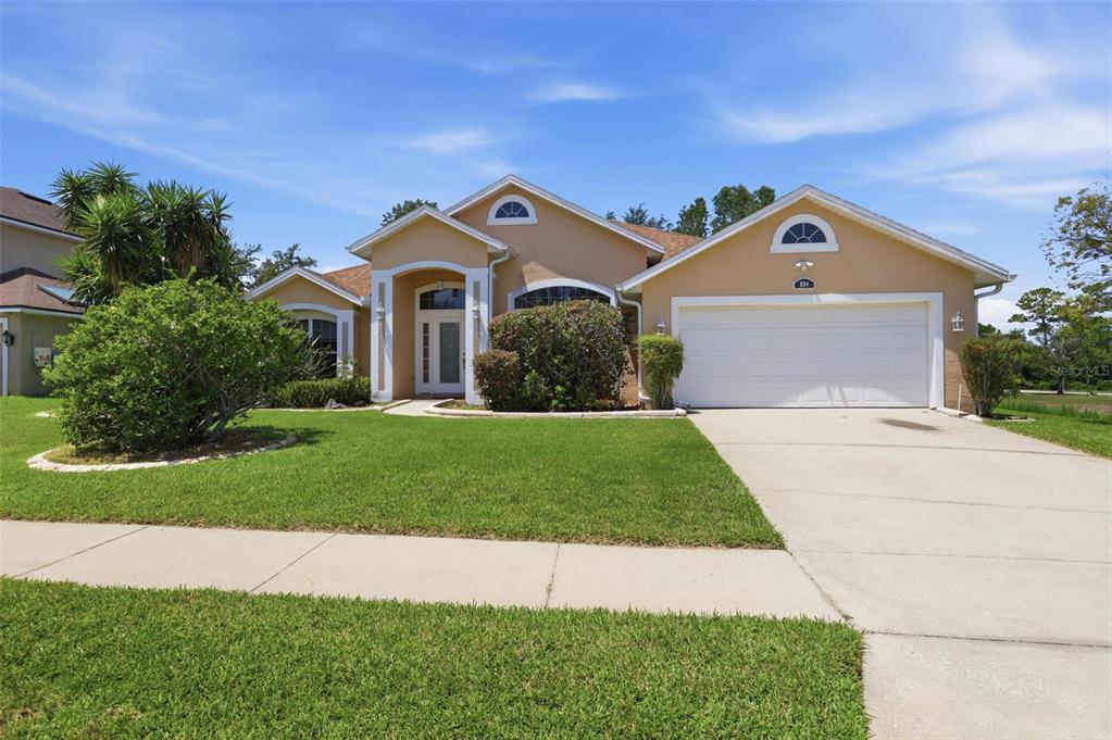 A single-story tan house with a two-car garage and a front lawn under a blue sky — 694 SHAWSBURY WAY, DELTONA, FL