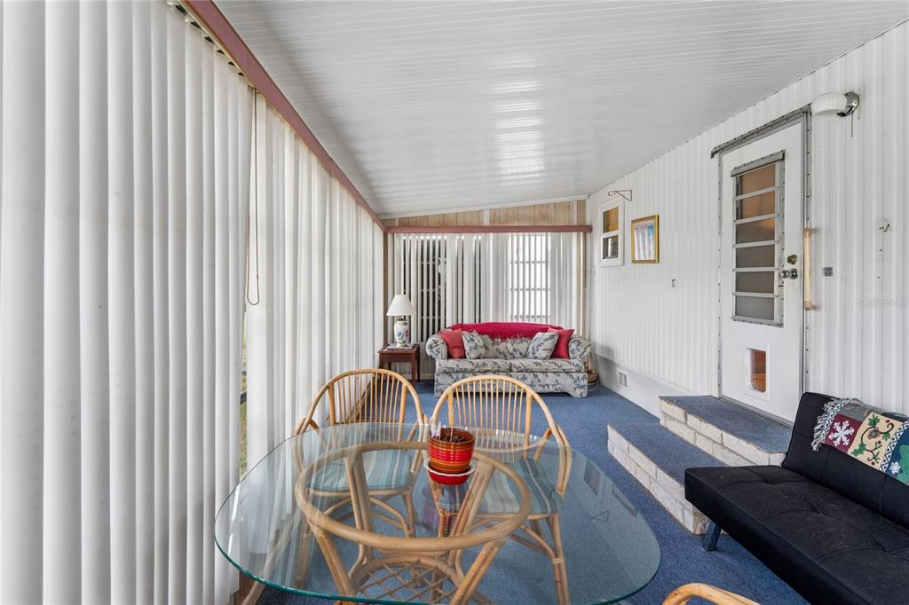 A sunroom features a glass dining table, blue carpeting, a sofa, and vertical blinds — 38618 REMORA AVENUE UNIT 92, ZEPHYRHILLS, FL