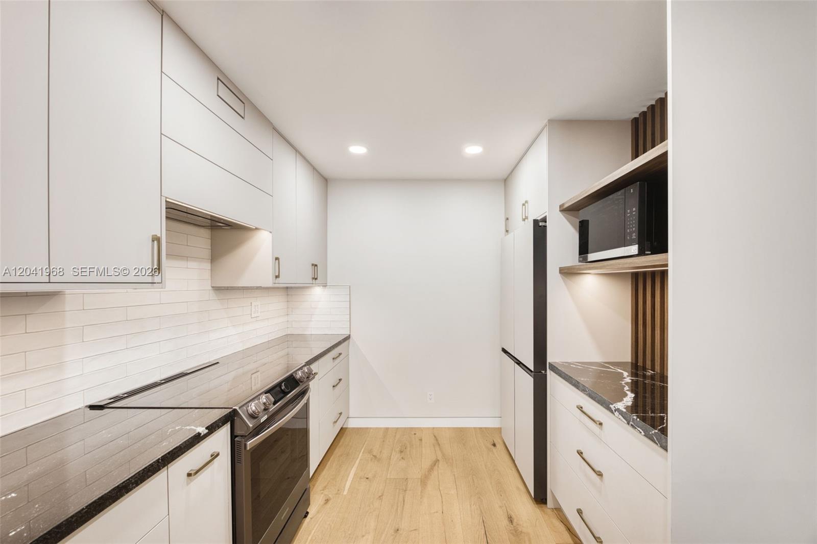 A modern galley kitchen features white cabinetry, black countertops, and light wood flooring — 100 Edgewater Dr # 142, Coral Gables FL 33133, CORAL GABLES, FL