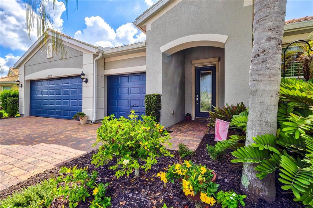 A single-story house exterior features a blue garage, a front entryway, and landscaped garden beds — 19556 PETRINO STREET, VENICE, FL