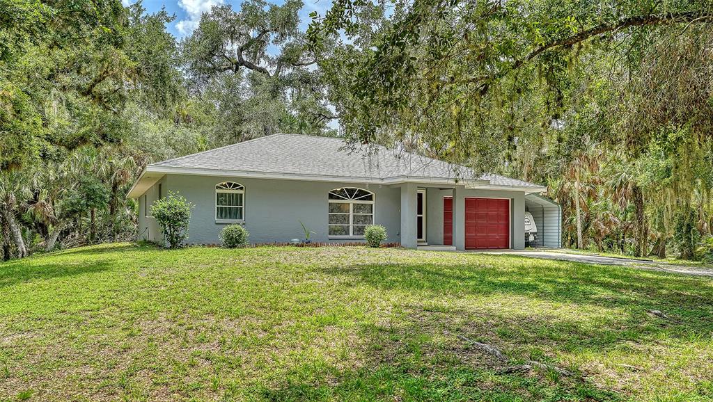 A gray single-story house with a red garage door sits on a grassy, wooded lot — 4498 HIDDEN RIVER ROAD, SARASOTA, FL