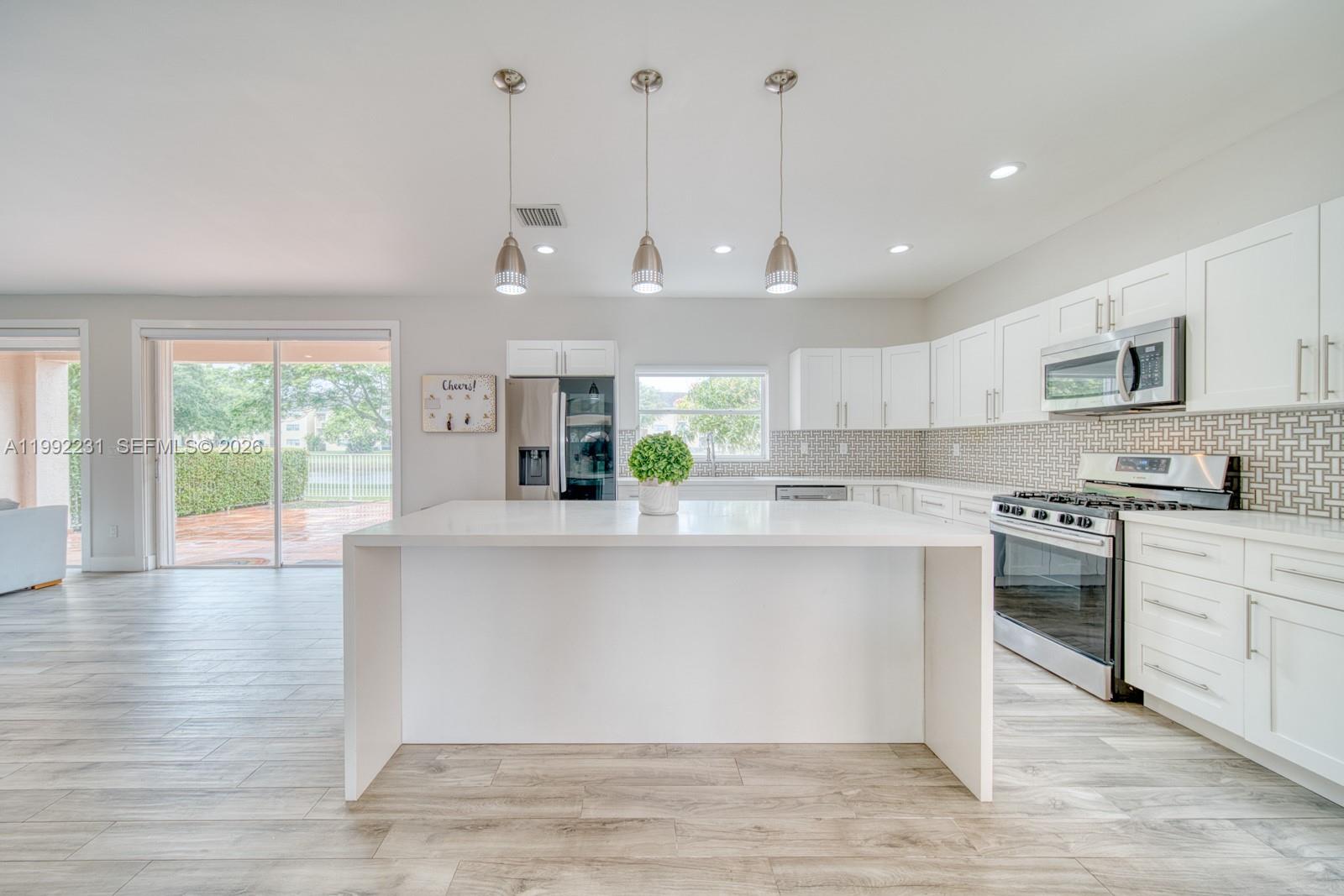 A modern white kitchen features a large center island, stainless appliances, and wood-look flooring — 15261 SW 108th Ter, Miami FL 33196, MIAMI, FL