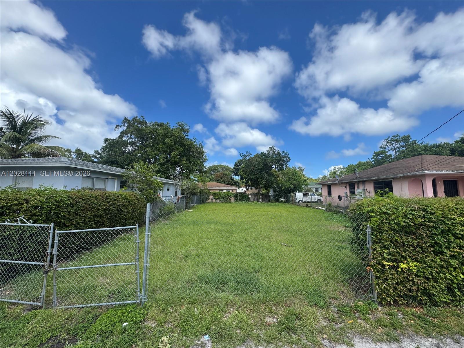 A fenced, grassy vacant lot sits between two residential houses under a blue sky — 6335 NW 1st Ct, Miami FL 33150, MIAMI, FL