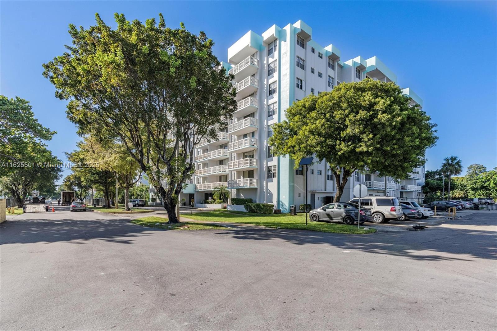 A multi-story apartment building with balconies stands behind a parking lot and large trees — 15600 NW 7th Ave # 306, Miami FL 33169, MIAMI, FL