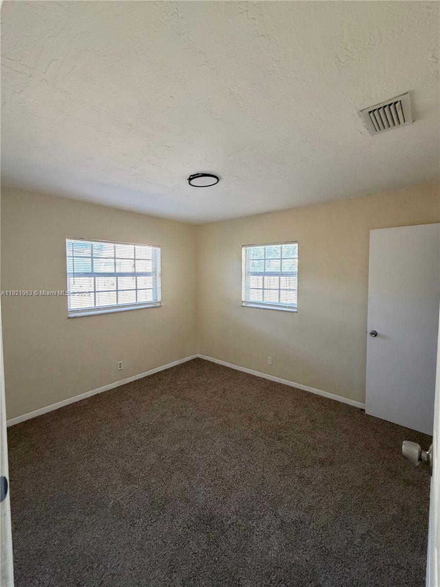 An empty bedroom features beige walls, brown carpeting, two windows, and a white door — 1320 SW 11th Ave # A, Deerfield Beach FL 33441, DEERFIELD BEACH, FL