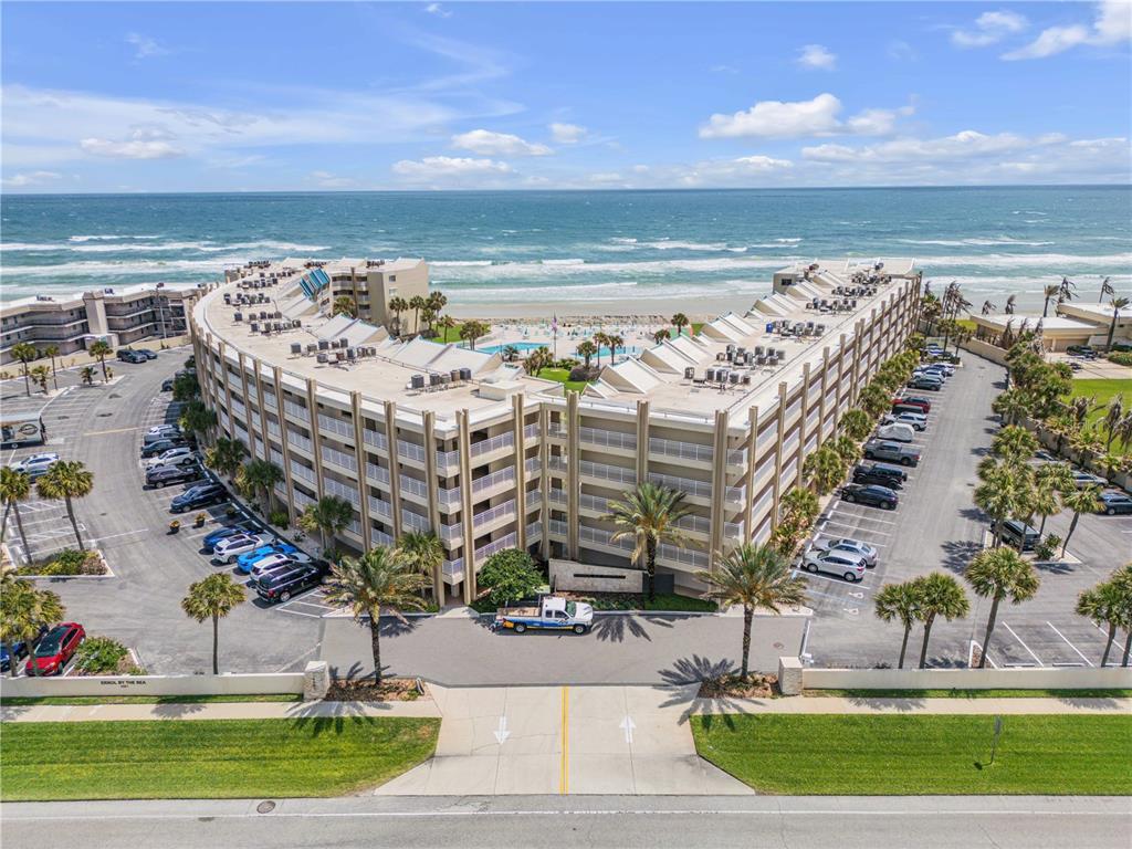 An aerial view shows a multi-story, curved beachfront condominium complex with parking lots and a pool — 4501 S ATLANTIC AVENUE UNIT 1150, NEW SMYRNA BEACH, FL