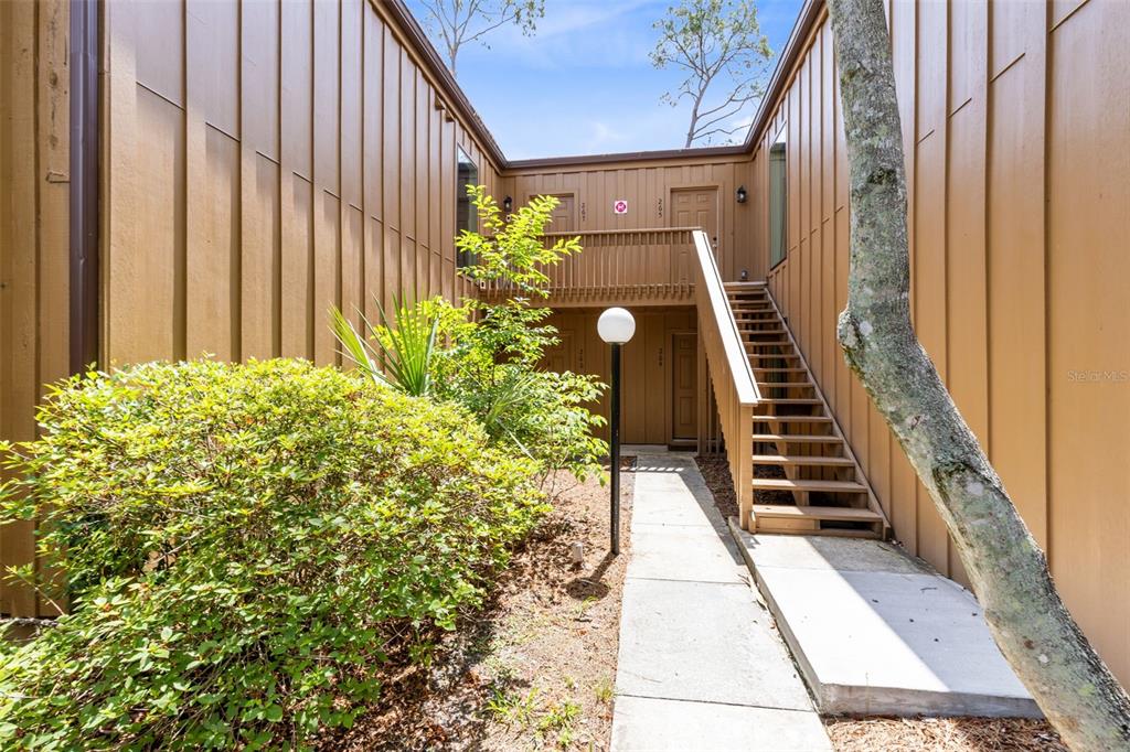 An outdoor walkway leads to a wooden staircase between two tan vertical-sided building sections — 267 CROWN OAKS WAY UNIT 267, LONGWOOD, FL