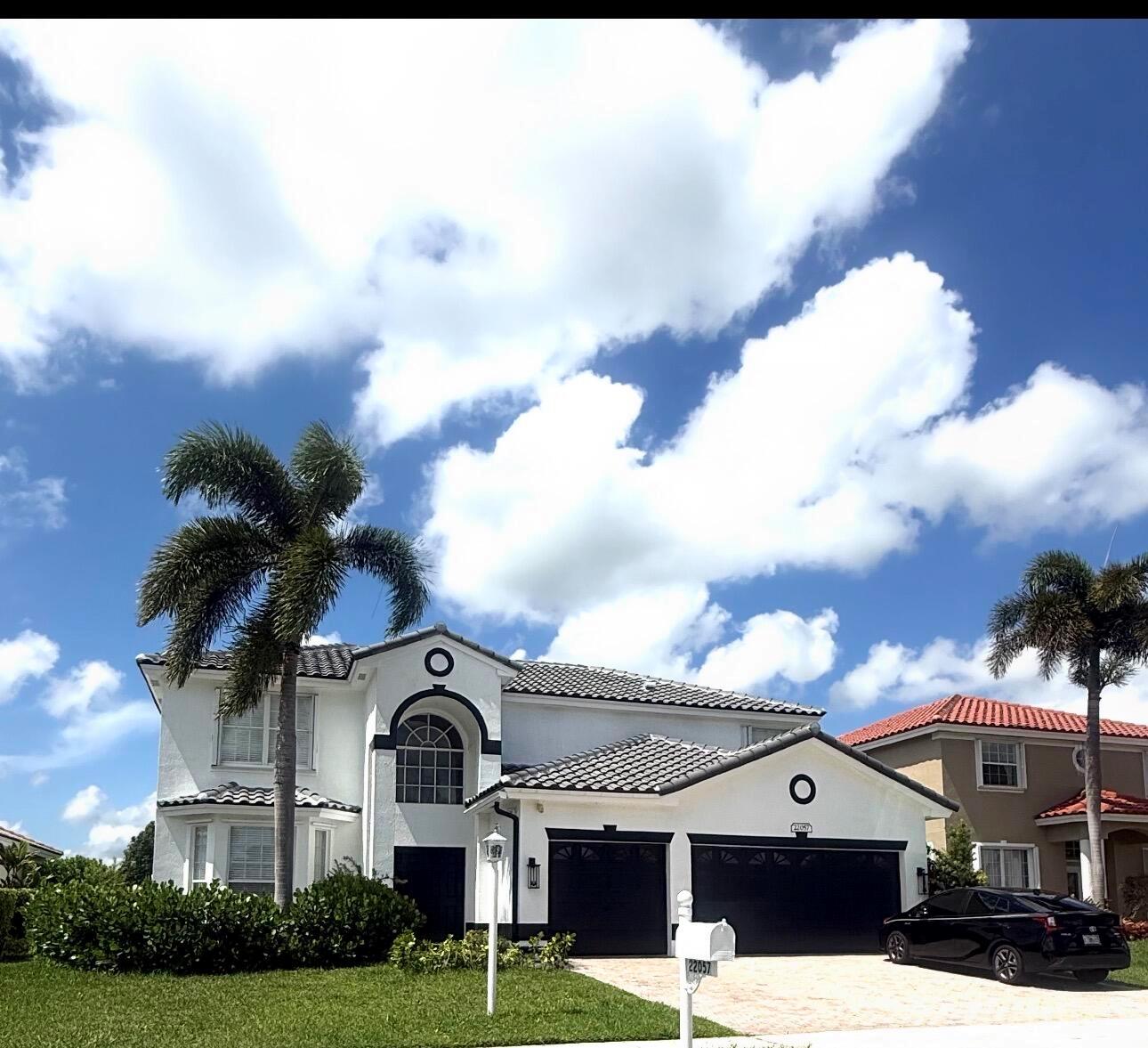 A two-story white house with a dark roof and garage doors sits under a cloudy sky — 22057 Palm Grass Drive, Boca Raton FL 33428, BOCA RATON, FL