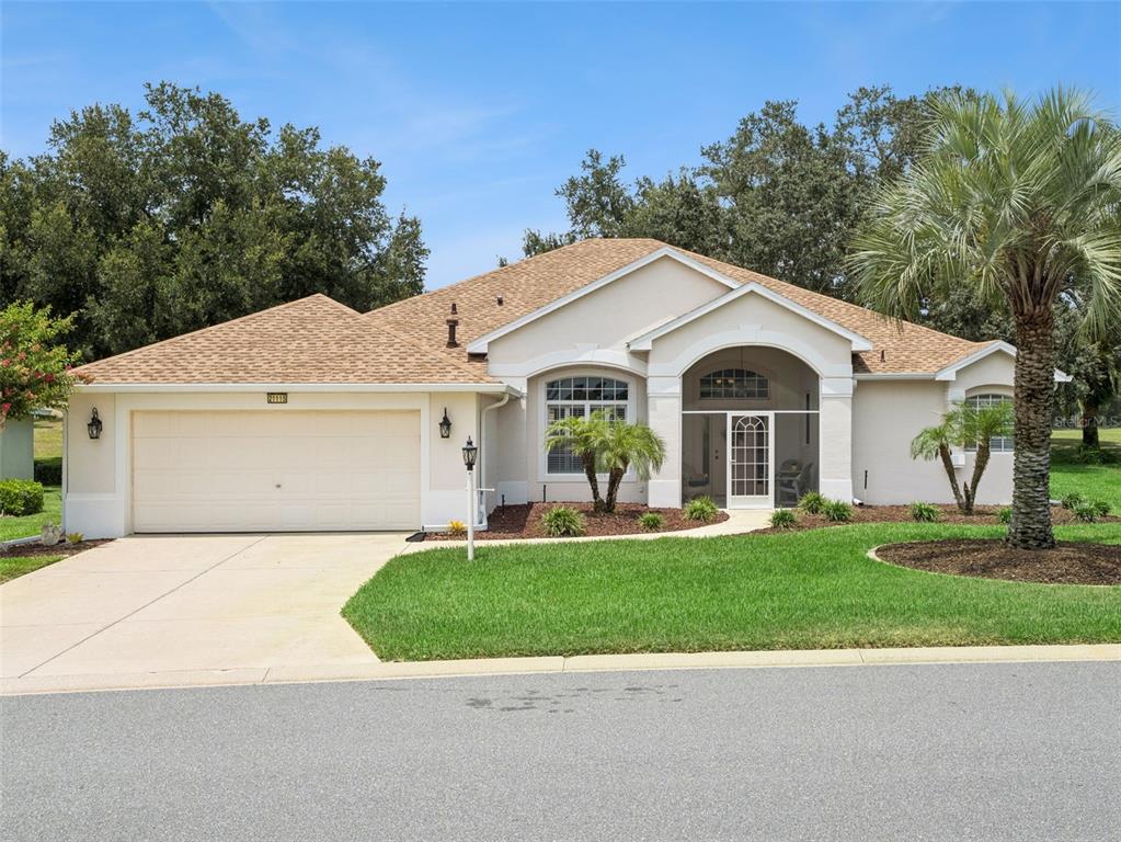 A single-story suburban house with a tan stucco exterior, a two-car garage, and a palm tree — 21115 ROYAL ST GEORGES LANE, LEESBURG, FL