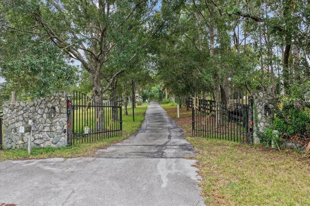 A paved driveway leads through an open metal gate flanked by stone pillars and trees — 470 FAY AVENUE, SPRING HILL, FL