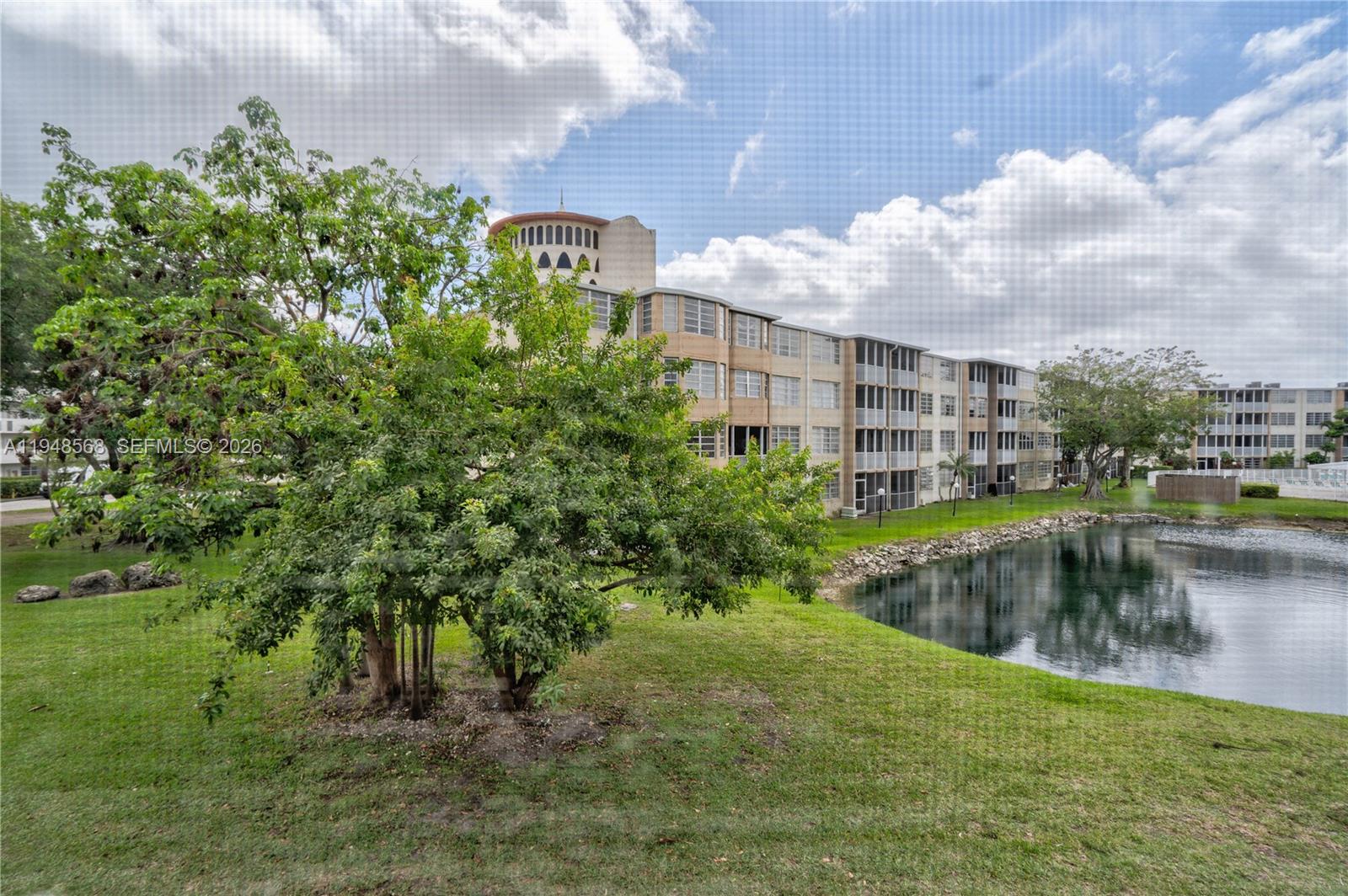 A multi-story apartment building stands behind a grassy lawn and a small pond — 1700 NE 191st St # 202, Miami FL 33179, MIAMI, FL