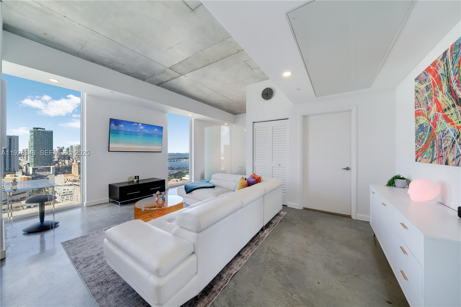 A modern living room features a white sectional sofa, concrete ceiling, and floor-to-ceiling city views — 151 SE 1st St # 3008, Miami FL 33131, MIAMI, FL