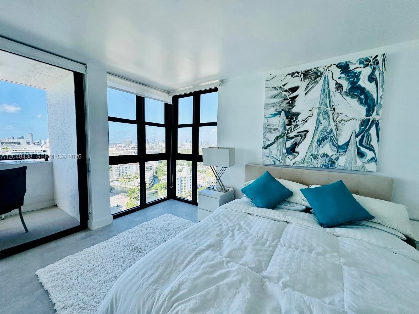 A bedroom features a bed with blue pillows, large windows with city views, and artwork — 2555 Collins Ave, Miami Beach FL 33140, MIAMI BEACH, FL