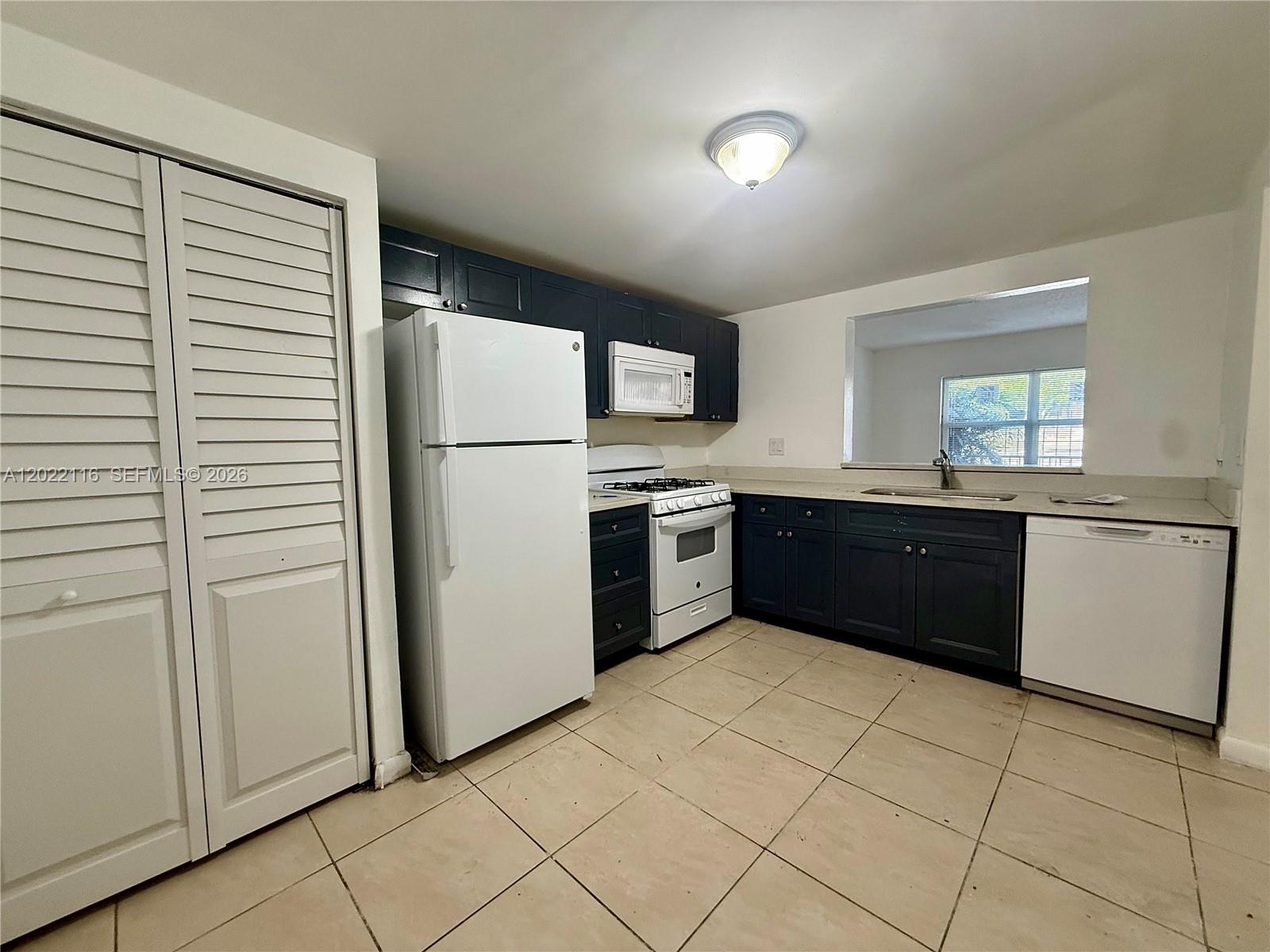 A kitchen features dark blue cabinets, white appliances, a pass-through window, and tiled flooring — 18101 NW 68th Ave # B102, Hialeah FL 33015, HIALEAH, FL