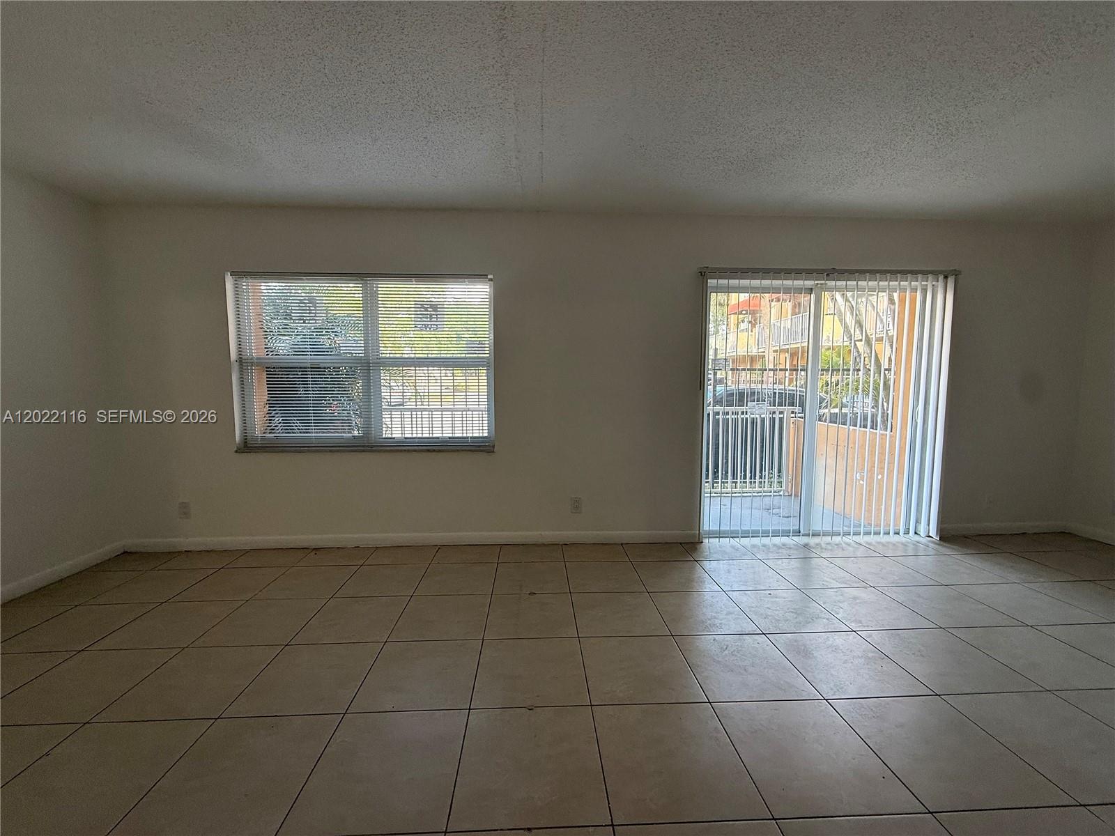 An empty room features beige tile flooring, a window with blinds, and a sliding glass door — 18101 NW 68th Ave # B102, Hialeah FL 33015, HIALEAH, FL
