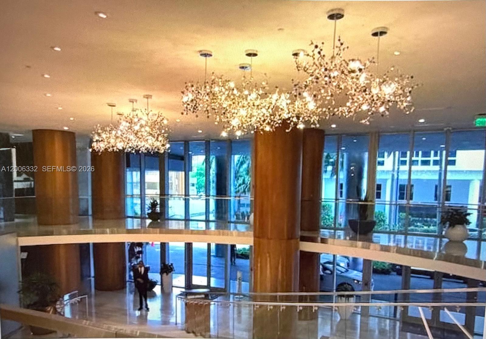 A modern building lobby features large wooden columns, glass walls, and ornate crystal chandeliers — MIAMI, FL