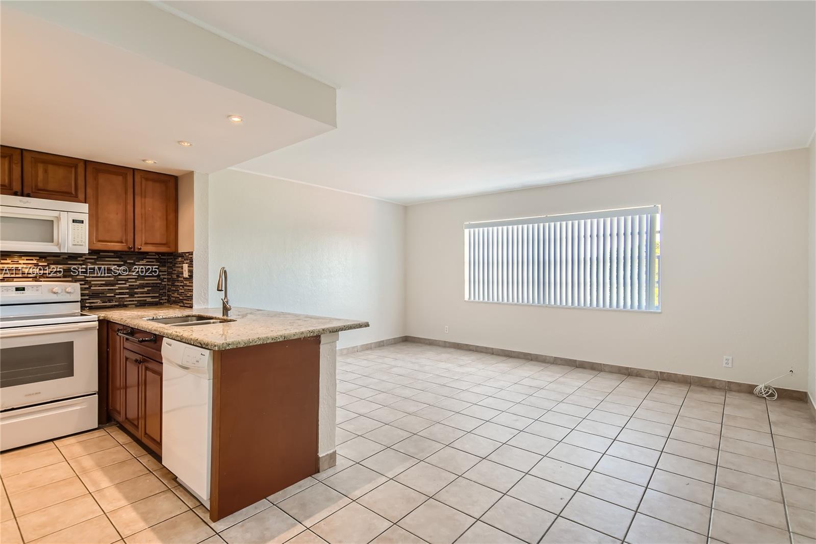 A kitchen with wood cabinets and a granite countertop opens to an empty tiled room — 1855 Plunkett St # 406, Hollywood FL 33020, HOLLYWOOD, FL