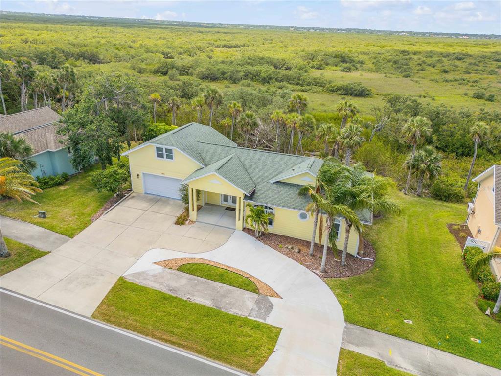 An aerial view shows a yellow house with a green roof, driveway, and surrounding vegetation — 1804 SAXON DRIVE, NEW SMYRNA BEACH, FL