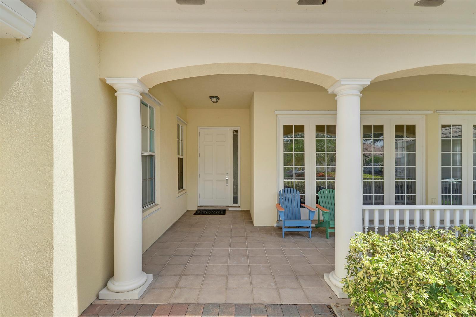 A covered porch features tiled flooring, white columns, a front door, and two Adirondack chairs — 8435 Belize Place, Wellington FL 33414, WELLINGTON, FL