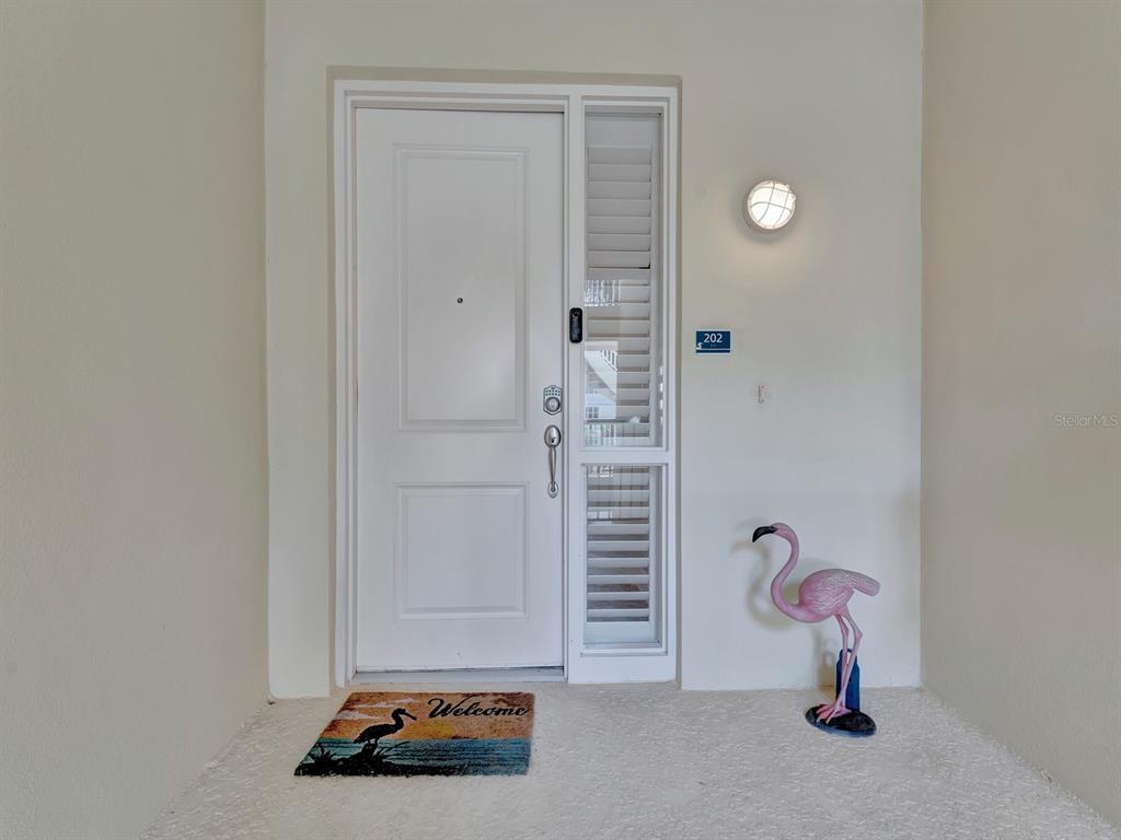 A white front door with a side window, a welcome mat, and a flamingo statue — 7840 34TH AVENUE W UNIT 202, BRADENTON, FL
