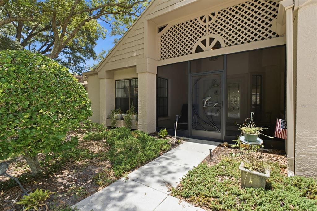 A concrete walkway leads to a screened-in entryway of a tan house with lattice detailing — 6695 PIN OAK COURT, SPRING HILL, FL