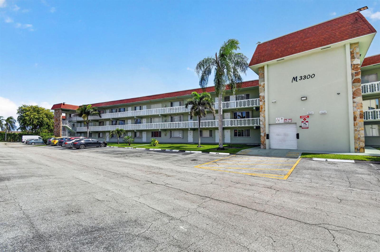 A three-story apartment building with a parking lot and palm trees in the foreground — 3300 Springdale Boulevard # 208, Palm Springs FL 33461, PALM SPRINGS, FL