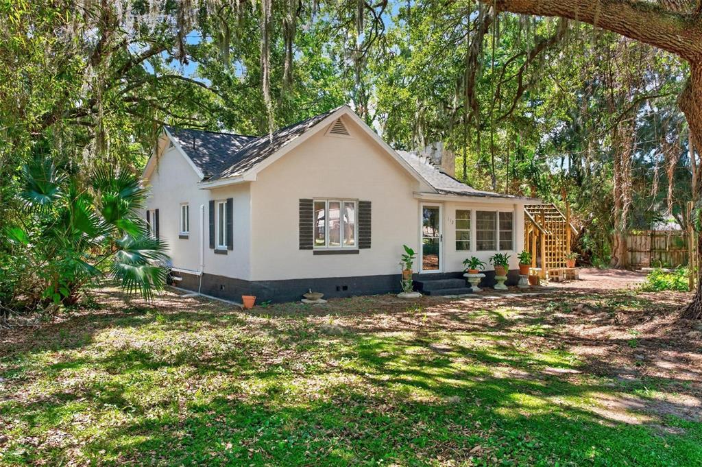 A single-story house with light-colored stucco walls and dark shutters sits under large oak trees — 112 N LAKE AVENUE, LEESBURG, FL