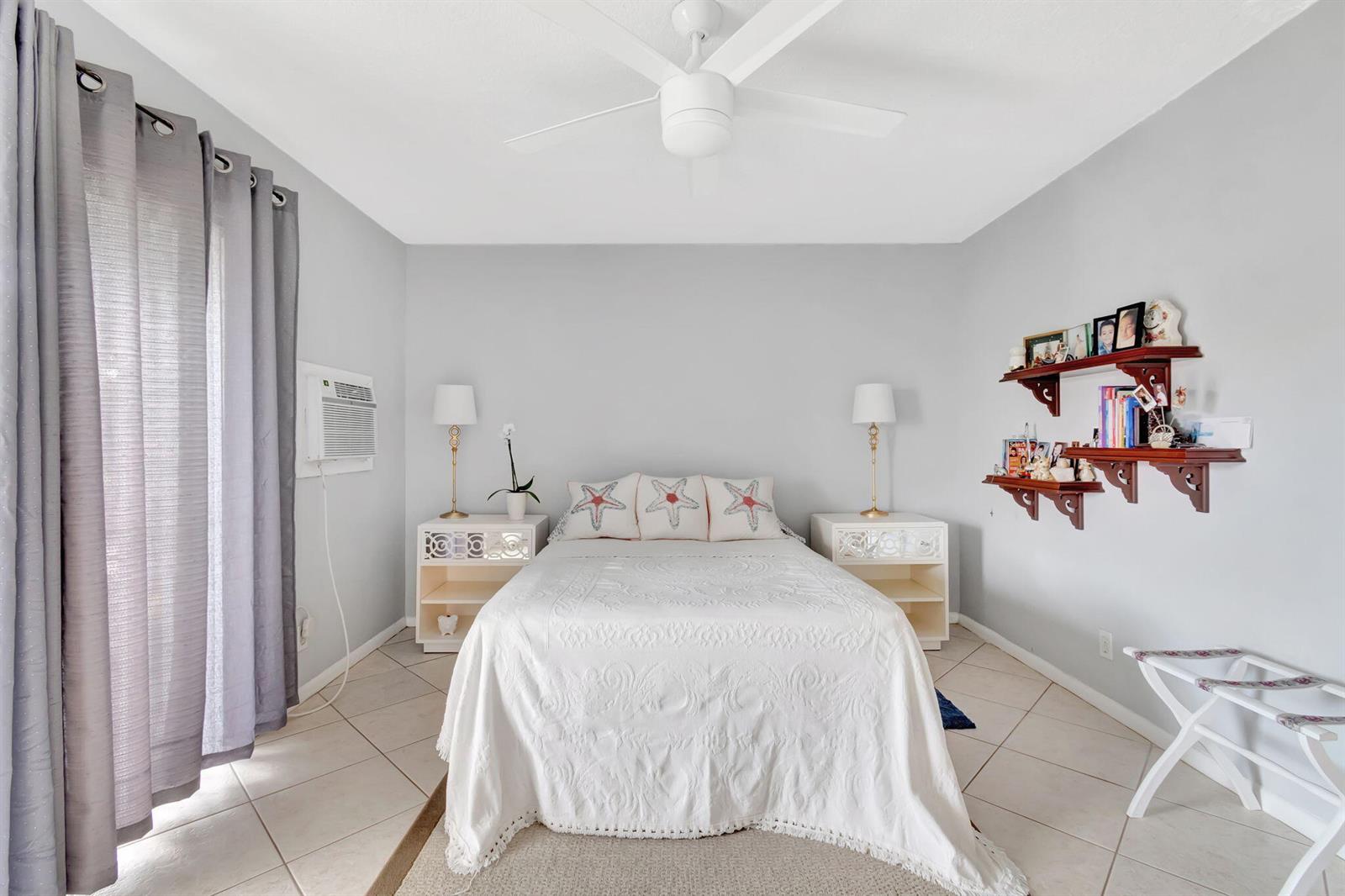 A bedroom features a bed with starfish pillows, two nightstands, wall shelving, and tile flooring — 46 Andover B, West Palm Beach FL 33417, WEST PALM BEACH, FL