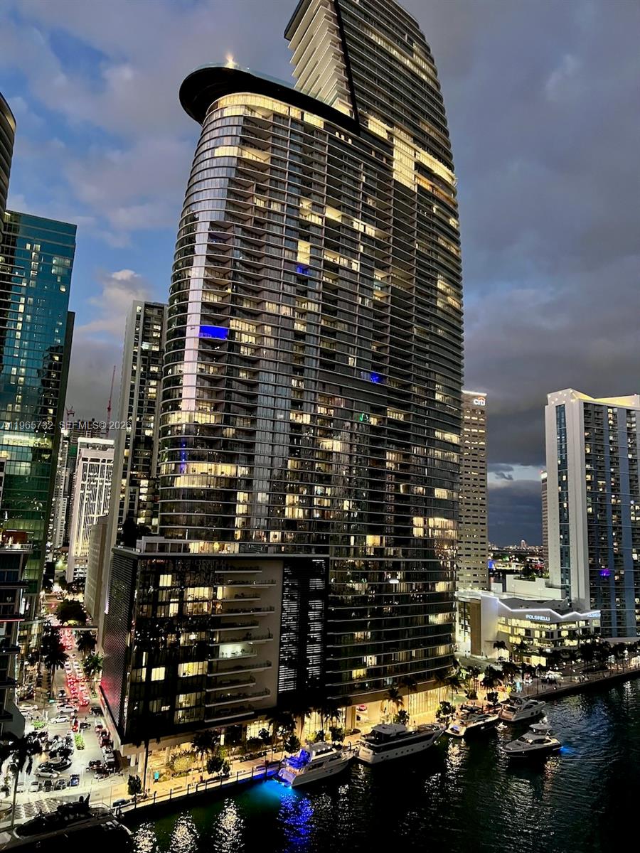 A tall, modern glass skyscraper stands illuminated at night beside a canal with docked yachts — 475 Brickell Ave # 1715, Miami FL 33131, MIAMI, FL