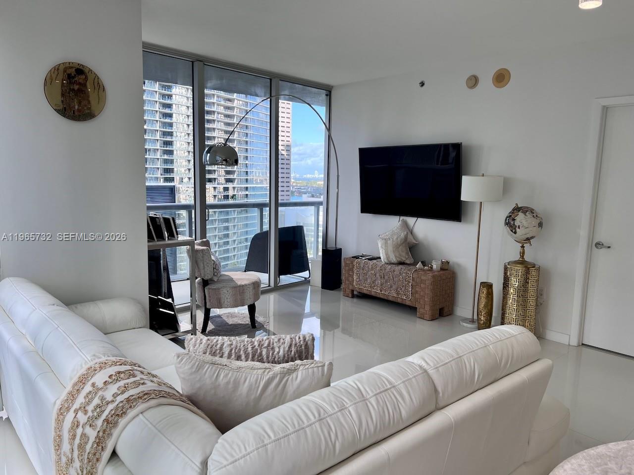 A modern living room features a white sofa, a wall-mounted television, and floor-to-ceiling windows — 475 Brickell Ave # 1715, Miami FL 33131, MIAMI, FL