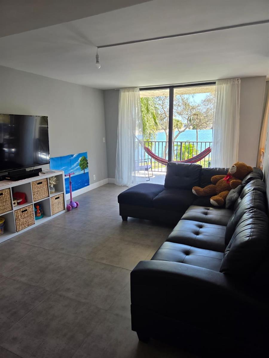 A living room with a black sectional sofa, media console, and a balcony overlooking water — 3500 Blue Lake Drive # 304, Pompano Beach FL 33064, POMPANO BEACH, FL