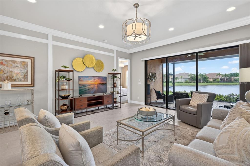 A living room features a television, shelving, gray furniture, and sliding doors overlooking a lake — 11957 FIVE WATERS CIRCLE, FORT MYERS, FL