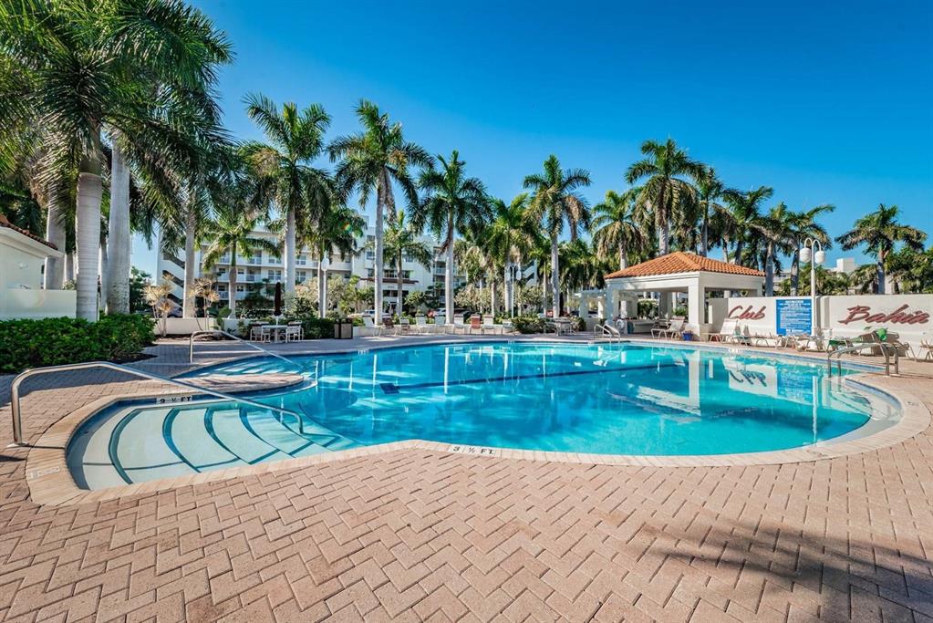 A swimming pool with steps and a surrounding brick patio features palm trees and buildings — 6083 BAHIA DEL MAR CIRCLE UNIT 460, ST PETERSBURG, FL