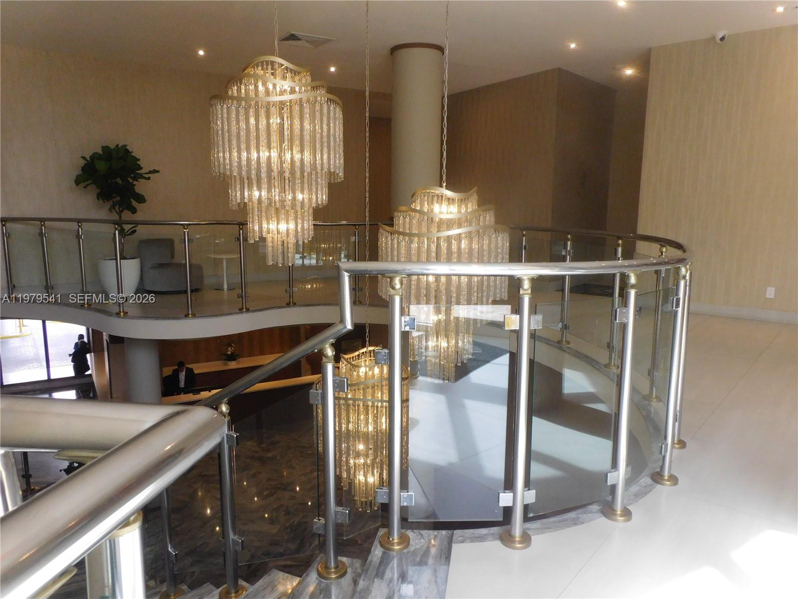 A curved staircase with a glass railing and three tiered crystal chandeliers in a lobby — 5401 Collins Ave # 526, Miami Beach FL 33140, MIAMI BEACH, FL