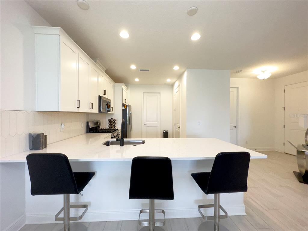 A modern kitchen features white cabinets, a quartz island with three black stools, and tile flooring — 17881 CHERISHED LOOP, BRADENTON, FL