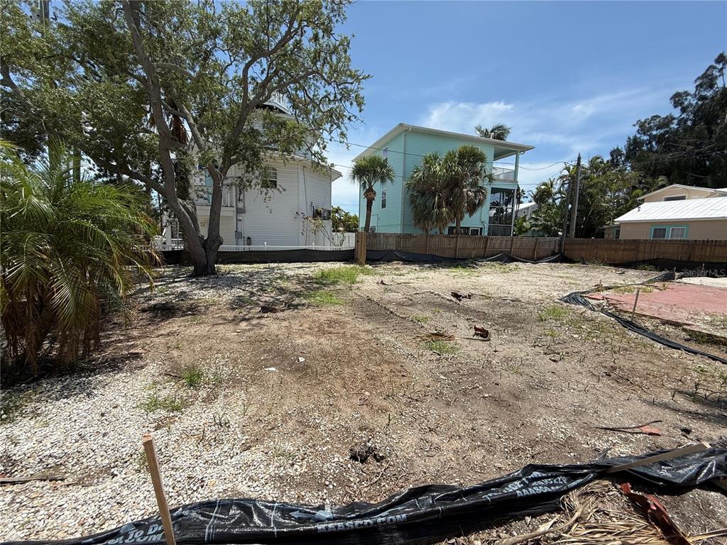A vacant, sandy residential lot is enclosed by a wooden fence with neighboring houses nearby — 8586 E BAY DRIVE, TREASURE ISLAND, FL