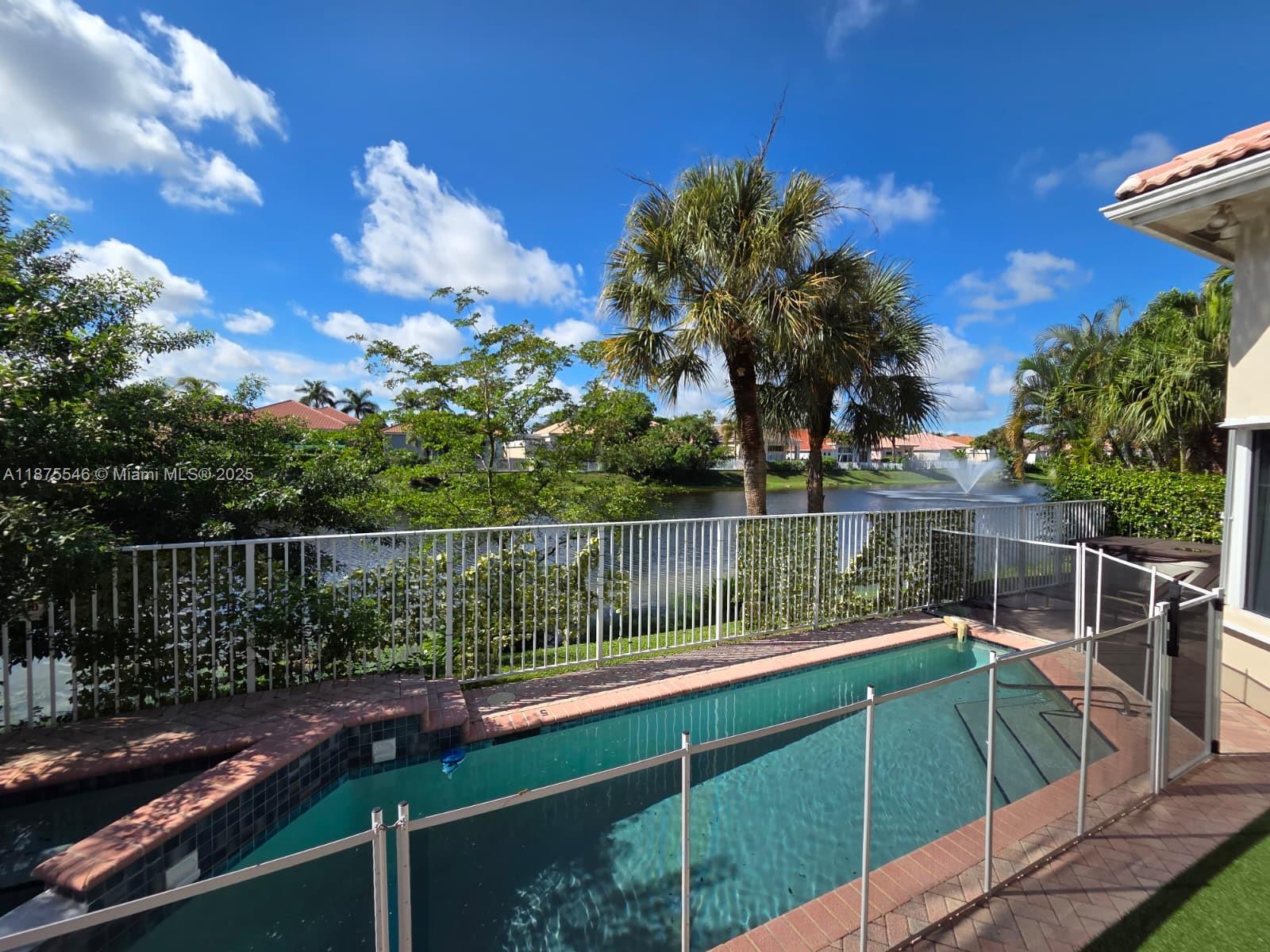 A swimming pool with a safety fence overlooks a lake with a fountain — 7030 Islegrove Pl, Boca Raton FL 33433, BOCA RATON, FL