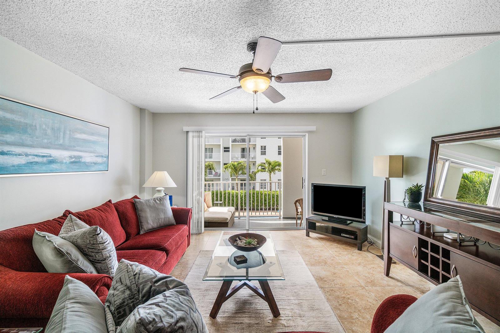 A living room features a red sofa, glass coffee table, ceiling fan, and balcony access — 950 Ponce De Leon Road # 209, Boca Raton FL 33432, BOCA RATON, FL