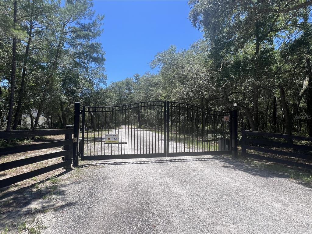 A black metal gate stands at the entrance of a gravel driveway lined with trees — 1425 RENBAR ROAD, DE LEON SPRINGS, FL