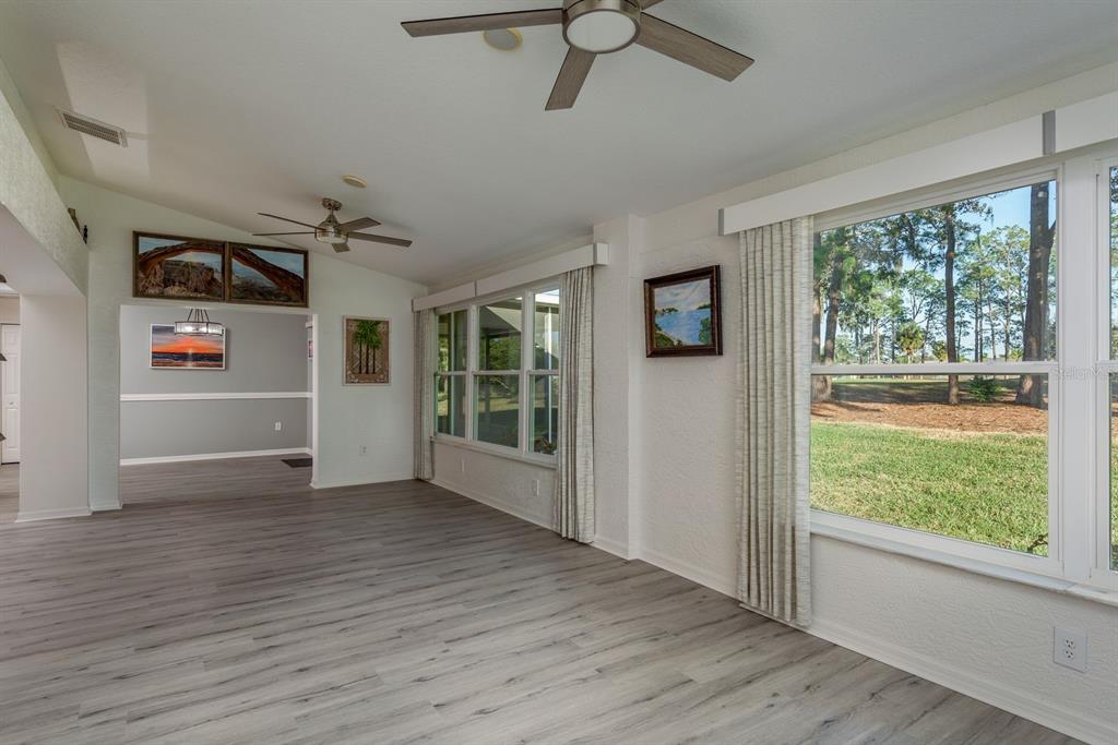 An empty living room features light wood-look flooring, ceiling fans, and large windows overlooking trees — 9742 OAKWOOD HILLS COURT, NEW PORT RICHEY, FL