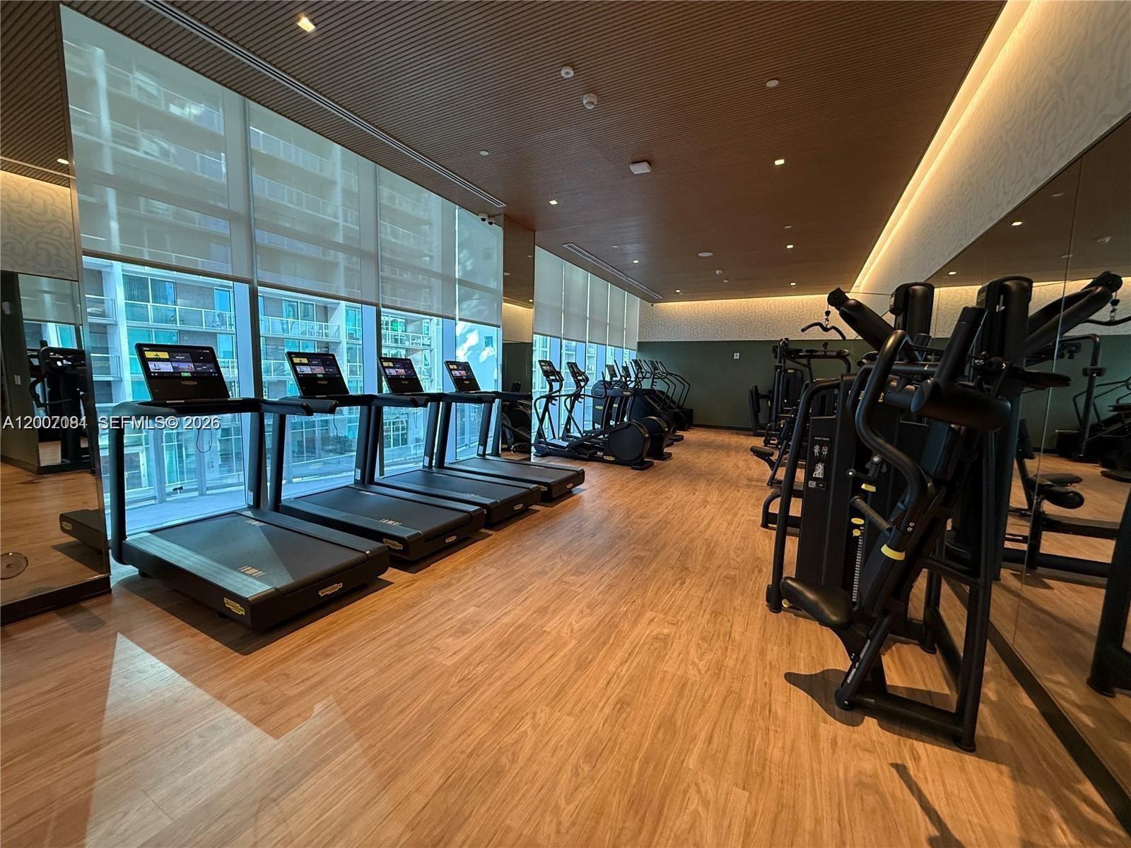 A modern gym features treadmills, weight machines, wood-look flooring, and floor-to-ceiling windows — 77 SE 5th Street # 1014, Miami FL 33131, MIAMI, FL
