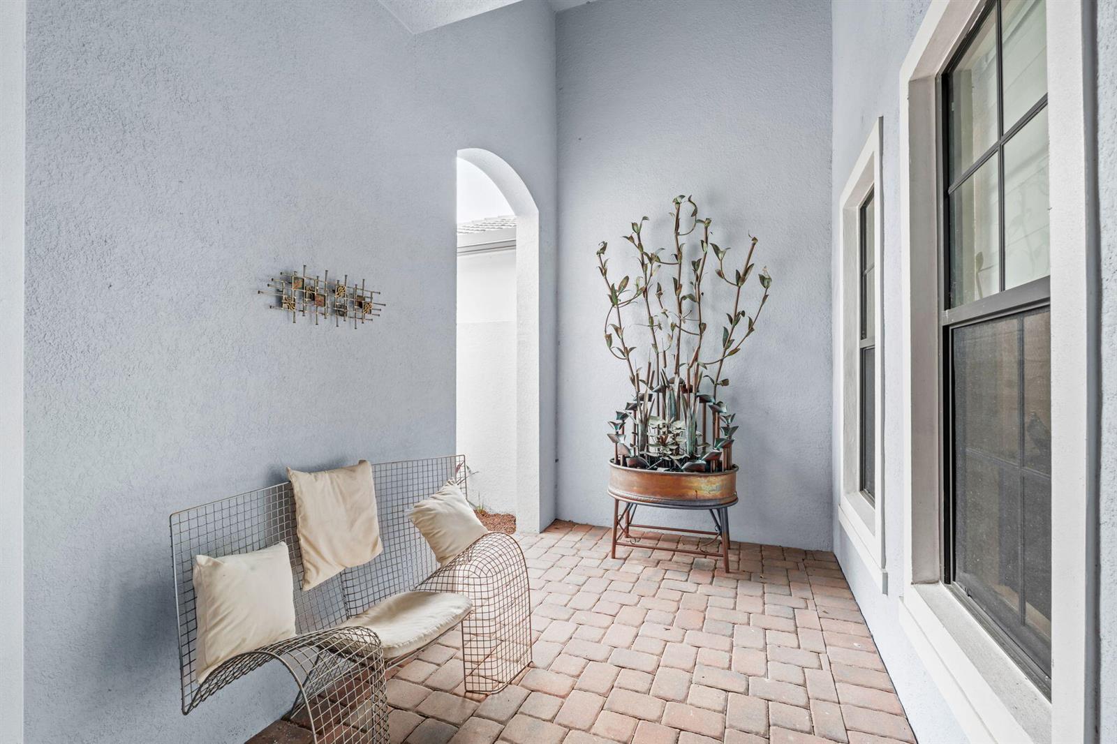 A covered brick patio features a wire-frame bench, a metal sculpture, and light blue walls — 7642 N Blue Spring Drive, Parkland FL 33067, PARKLAND, FL