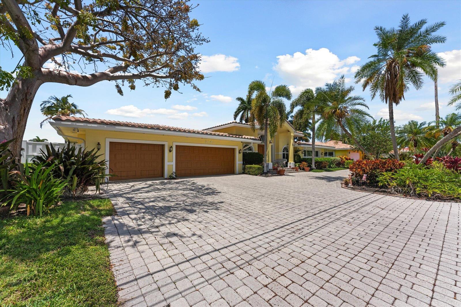 A yellow house with a two-car garage and a large paver driveway under palm trees — 2141 NE 29th Street, Lighthouse Point FL 33064, LIGHTHOUSE POINT, FL