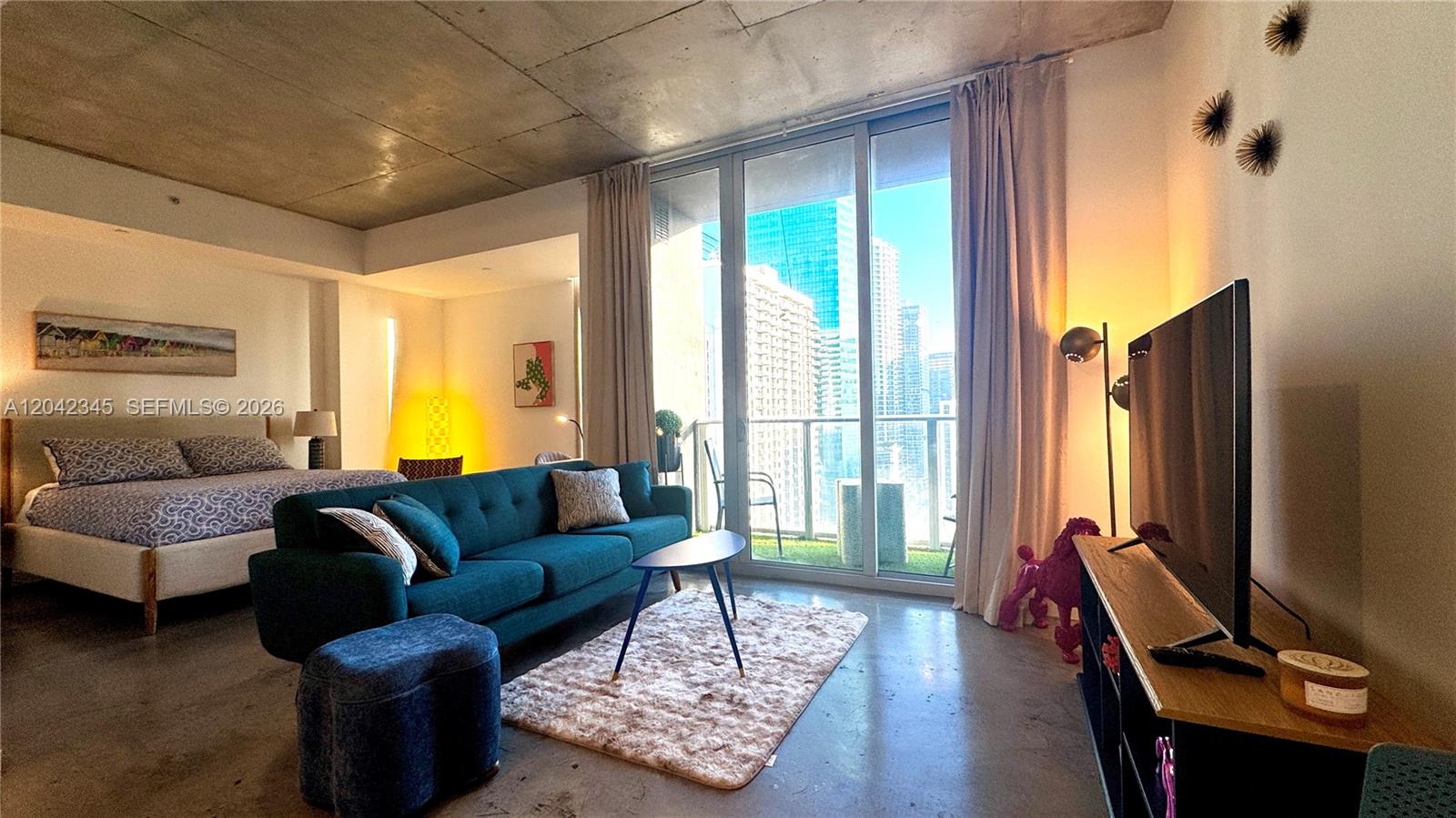 A studio apartment features a teal sofa, a bed, and floor-to-ceiling windows with city views — 151 SE 1st St # 2205, Miami FL 33131, MIAMI, FL